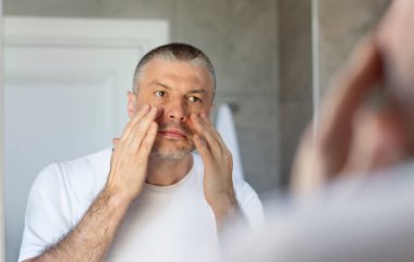 Sıkıcı cilt. Orta yaşlı bir adam banyoda aynaya bakıyor ve yüzüne dokunuyor, gözlerinin altında siyah halkalar var, görünüşünden memnun değil.