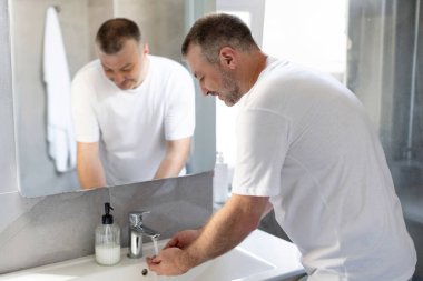 Adam ellerini yıkıyor, lavabonun yanında duruyor, evdeki tuvalette sabah temizliği yapıyor.