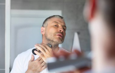 Orta yaşlı Avrupalı bir adam tıraş makinesi ya da elektrikli traş makinesiyle sakal tıraşı olurken aynadaki yansımasına bakıyor.