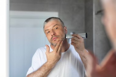 Adamın yansıması aynanın önünde duruyor ve sabah banyoda sakalını tıraş etmek için tıraş makinesi kullanıyor.