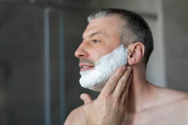 Erkeklere özel bakım konsepti. Yakışıklı bir adam yüzüne tıraş köpüğü sürüyor, sabahları tıraş olmaya hazırlanıyor.