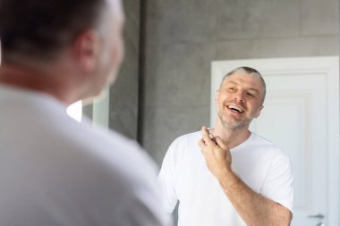 Mutlu Avrupalı orta yaşlı adam aynaya bakıyor, parfüm sıkıyor, sabah cilt bakımı yapıyor. Nemlendirici ürünler ve parfüm