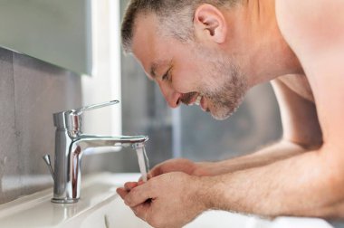 Erkek sabah rutini. Çıplak gövdeli bir adam yüzünü suyla yıkıyor lavabonun yanında duruyor musluk başında duruyor, günlük rutin şeyler yapıyor, yandan bakıyor.