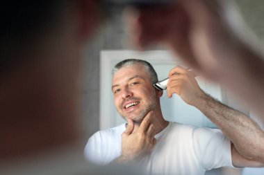 Yakışıklı, orta yaşlı bir adam banyoda aynanın karşısında duruyor ve sakalını tıraş ediyor.