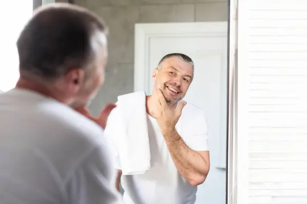 stock image Handsome European middle aged man in t-shirt looking in mirror in bathroom and touching chin, smiling to his reflection