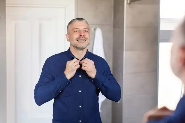 stock image Excited middle aged businessman getting ready and buttoning up blue shirt, standing in bathroom in front of mirror, preparing to go to work