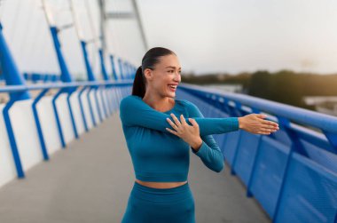 Açık havada antrenman yapmadan önce gülümseyen, formda genç bir kadın, köprüde koşmaya hazırlanıyor, kollarını açıyor, yan tarafa bakıyor ve gülümsüyor.