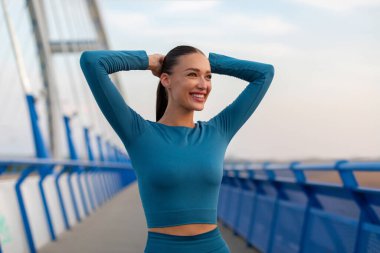 Çekici genç Avrupalı kadın koşmadan önce egzersiz yapıyor, ısınıyor ve gülümsüyor, akşamları köprüde duruyor.