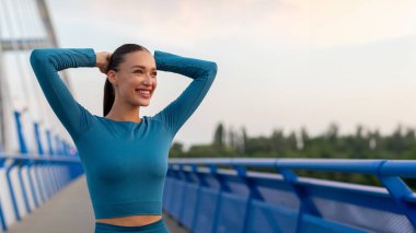 Spor giyim sektöründe genç ve mutlu bir kadın. Akşamki antrenman sırasında köprüde ellerini uzatıyor. Panorama, serbest alan.
