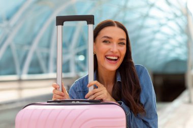 Havaalanı terminalinde bekleyen heyecanlı genç Avrupalı kadın, bavuluyla oturuyor ve kameraya gülümsüyor, seyahatin tadını çıkarıyor.