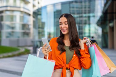 Alışverişkolik kadın cep telefonu kullanıyor ve alışveriş yaptıktan sonra elinde renkli kağıt torbalarla dışarıda bekliyor. Ticaret konsepti sunar