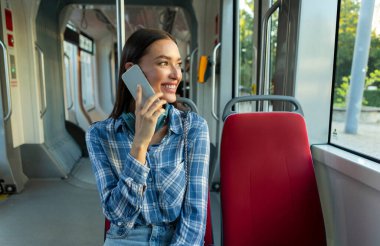 Devlet otobüsünde ya da tramvayda otururken cep telefonuyla konuşan gülümseyen genç bayan, koltukta oturan ve pencereden dışarı bakan kadın yolcu.