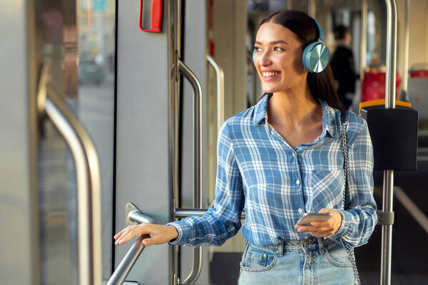 Молодая женщина, стоящая в общественном транспорте, современный трамвай, улыбающаяся, слушающая музыку в наушниках и смотрящая в окно. Общественный транспорт, городской образ жизни