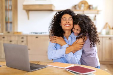 Mutlu anne dizüstü bilgisayarda çalışırken kızı arkadan annesini kucaklıyor, kadın evde mutfak masasında oturuyor.