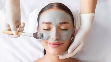 Kozmetik uzmanı, lüks kaplıca salonundaki genç bir kadına cilt bakımı yapıyor, rahat bir bayanın yüzüne kilden maske takıyor.