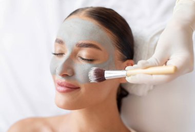 Güzellik salonundaki güzellik uzmanı genç bir kadına gri doğal kilden yüz maskesi takıyor. Sağlık ve cilt bakımından zevk alan bir bayan. Yakın plan.