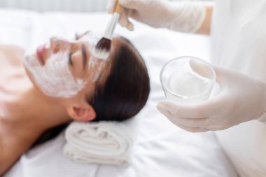 Rahatlamış genç bir kadın güzellik salonunda yüz bakımı yaptırıyor. Güzellik uzmanı alnına kille dokunuyor. Kaseyle eline odaklanıyor.