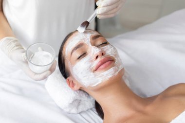 Spa merkezinde cilt bakımı gören genç bir kadın, profesyonel kozmetik uzmanı güzel bir bayanın yüzüne nemlendirici maske takıyor.