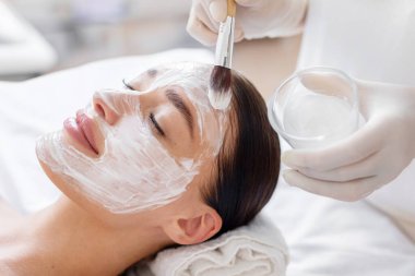 Cilt bakımı. Spa salonunda kozmetik maske takan bir kadın, kozmetik uzmanı elleri kadın yüzüne fırça sürerek maske takıyor.