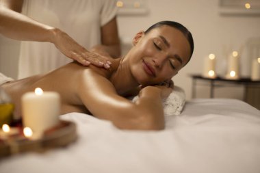 Rahatlamış genç bir kadın, yanan mumlarla süslenmiş lüks kaplıcalarda aromaterapi ile iyileştirici vücut masajı yaptırıyor.