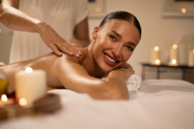 Spa salonundaki masajda uzanmış gülümseyen kadın, terapist omuzlarını ve sırtını sıvazlarken kameraya gülümsüyor.