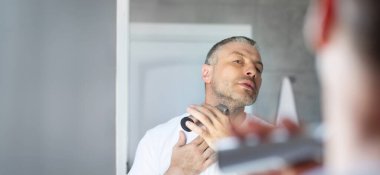 Orta yaşlı Avrupalı bir adam tıraş makinesi ya da elektrikli traş makinesiyle sakal tıraşı olurken aynadaki yansımasına bakıyor.