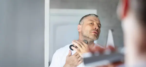 stock image Middle aged European man shaving beard with trimmer or electric shaver, looking at his reflection in mirror, at bathroom interior