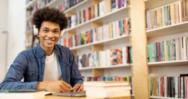Kolej kütüphanesinin iç kısmında, kulaklık takan ve dizüstü bilgisayar önünde notlar alan mutlu siyahi öğrencinin portresi.