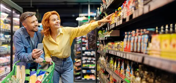 Çift birlikte market alışverişi yapıyor, erkek cep telefonu kullanıyor, kadın rafı işaret ediyor, süpermarketten yiyecek ürünleri seçiyor.