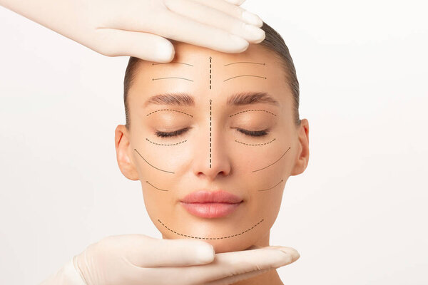 Portrait of young woman getting beauty treatment from beautician in protective gloves, lady with arrows on face isolated on white studio background