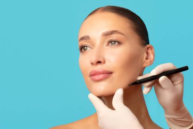 Estetik cerrah ameliyattan önce yüz hatlarını siyah kalemle çiziyor, genç bayan estetik ameliyat oluyor, mavi arka plan, boş alan.