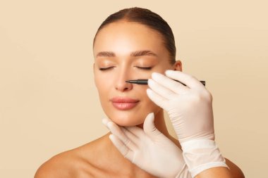 Yüz estetiği konsepti. Doktor cerrahın elinde kalem var ve danışma sırasında kadının yüzüne işaret koymaya hazır, bej rengi stüdyo arka planı.