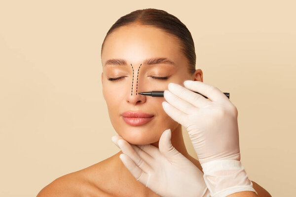 Doctor making marks on female nose, young woman posing on beige studio background with closed eyes during consultation. Rhinoplasty concept.