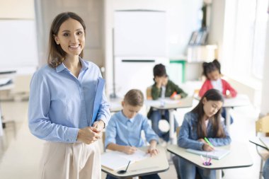 İlkokulda öğrencilerin sınıfta arka plan üzerine kitap yazarken kameraya gülümseyen mutlu arkadaş canlısı bayan öğretmen.