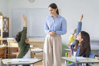 Mutlu okul çocukları ders sırasında cevap vermek için ellerini kaldırıyor. Dost canlısı bayan öğretmen çocuklara bakıyor ve gülümsüyor.