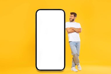 Havalı bir adam dev cep telefonuna yaslanmış, kollarını kavuşturmuş ve beyaz ekran sarı stüdyo arka planına bakıyor, mobil uygulama ya da reklam için modelleme yapıyor.
