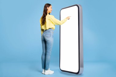 Gündelik kıyafetli kadın büyük akıllı telefonun önünde duruyor, boş ekrana dokunuyor, en yeni mobil uygulama ya da web sitesini kullanıyor, mavi arka plan, model