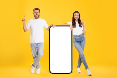 Avrupalı çift boş ekranlı büyük akıllı telefonun yanında duruyor, yukarı bakıyor, sarı arka planda poz veriyor, tasarım için model, reklam şablonu
