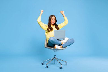 Dizüstü bilgisayarlı koltukta oturan heyecanlı Avrupalı bayan mavi arka plandaki başarısını kutluyor. Büyük satışlardan hoşlanan kadınlar veya online kazananlar