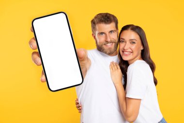 Cep telefonu reklamı. Kameraya boş akıllı telefon gösteren mutlu Avrupalı çift, sarı stüdyo arka planında fotokopi alanını kucaklıyor ve gösteriyorlar.
