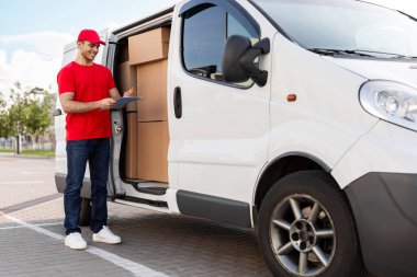 Üniformalı genç bir kurye müşteri adresleriyle panosunu kontrol ediyor. Paketlerle dolu, uzun menzilli bir minibüsün yanında duruyor.