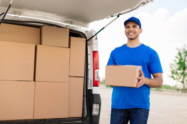 Posta servisi konsepti. Üniformalı genç teslimatçı elinde paket kutusu, gülümsüyor, dışarıda minibüsün yanında duruyor.