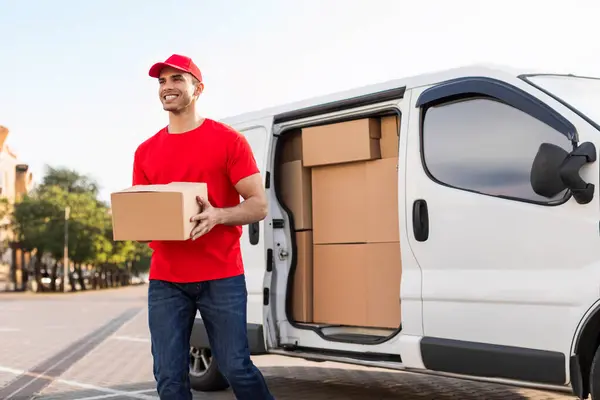 stock image Cheerful young delivery man in red uniform carrying package cardboard box from his white van, delivering parcel to customer