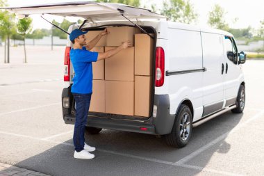 Üniformalı kurye çeşitli karton kutuları minibüse yüklüyor, paket teslimatına hazırlanıyor, otoparkta arabanın yanında duruyor.