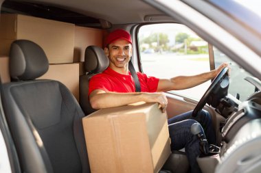 Hızlı teslimat. Paketler ve kutularla dolu bir minibüste oturan mutlu kurye kameraya bakıp gülümsüyor.