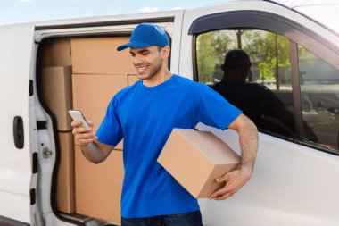 Paket kutusunu tutan ve cep telefonu kullanan mutlu teslimatçı dışarıda minibüsün yanında duruyor ve müşteriyi bekliyor.