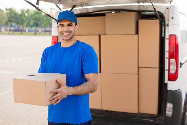 Young European man holding cardboard box parcel, delivering it to customer home address, guy looking and smiling at camera