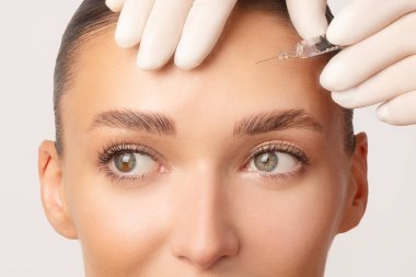 Avrupalı genç bir kadının beyaz arka plan üzerinden alnına güzellik iğnesi vurulduğu vesikalık fotoğraf. Elinde şırınga tutan koruyucu eldivenli doktor.