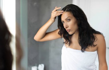 Saçlarının köklerine bakan üzgün genç bir bayan. Duştan sonra havluya sarılmış, kuru, pullu kafa derisinden acı çekiyor.