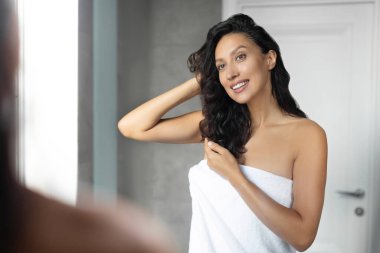 Güzel Avrupalı bayan kıvırcık saçlarıyla ilgileniyor, aynanın önünde duruyor ve gülümsüyor.